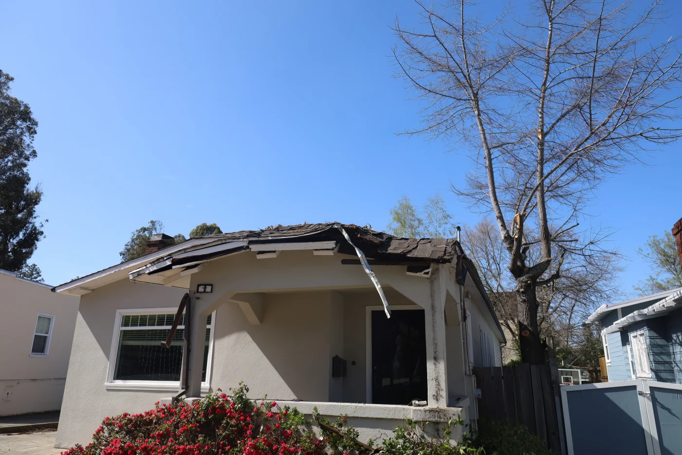 Image of a small house with bushes in front of the porch and a damaged, bent roof