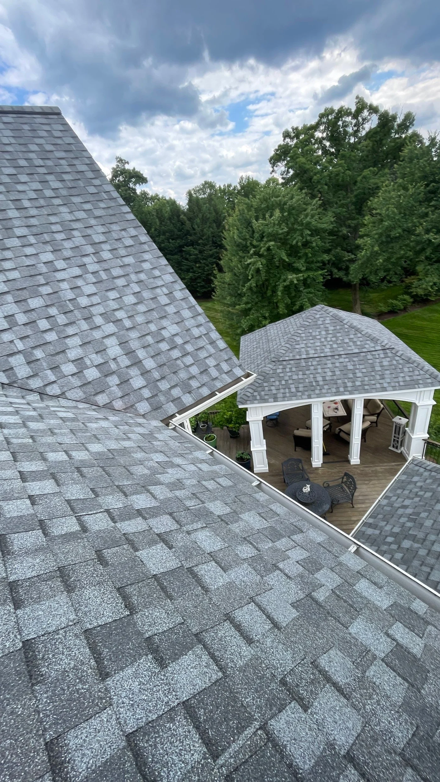 A top-down edge view of a light-grey shingle roof looking down into an outdoor seating area