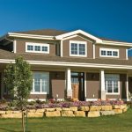 Brown home with white-trimmed windows, covered porch, stone columns, shingle roof, garden bed, rock wall, green lawn, and blue sky.