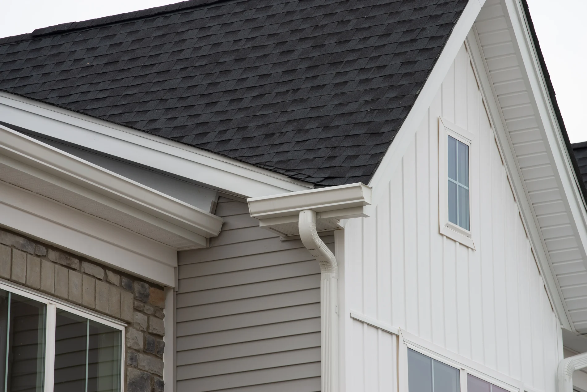 A close-up shot of one section of a roof covered in black asphalt shingles. It shows where two slopes meet. The house has tan siding and white gutters and trim.