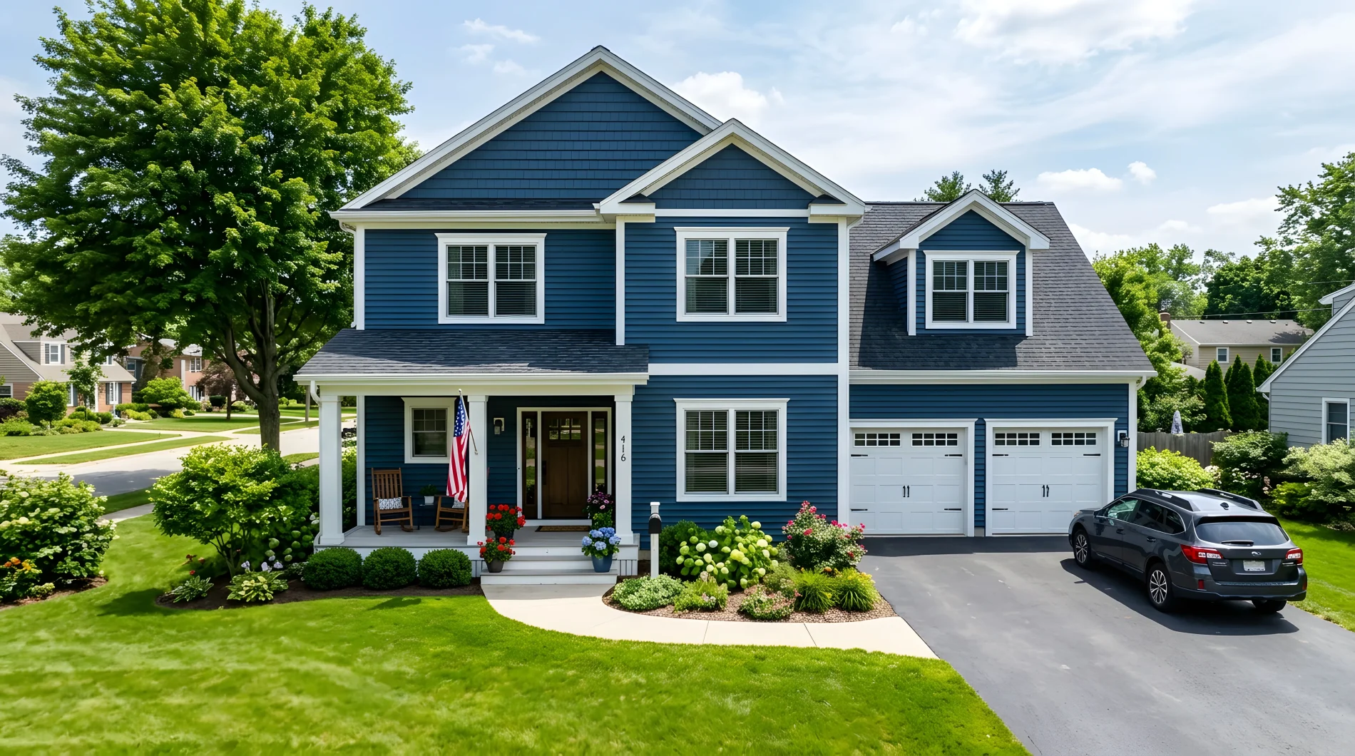 A multi-story home with dark blue siding, white trim and window frames, and a dark gray shingle roof. The home has a covered front porch on the left side, and a two-car garage on the right.