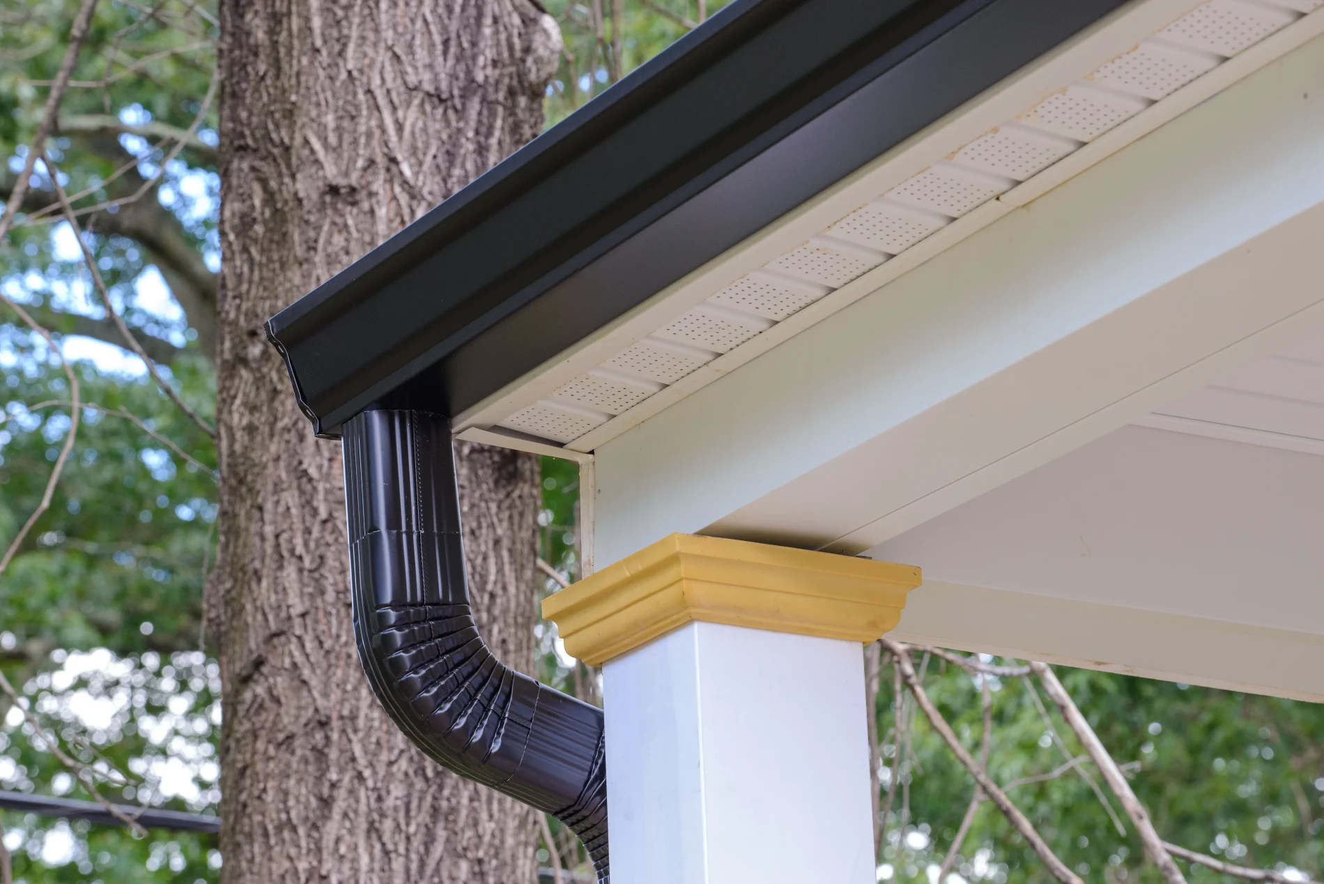 The front corner of a home’s covered porch, showing white soffit and fascia and a black metal gutter and downspout.