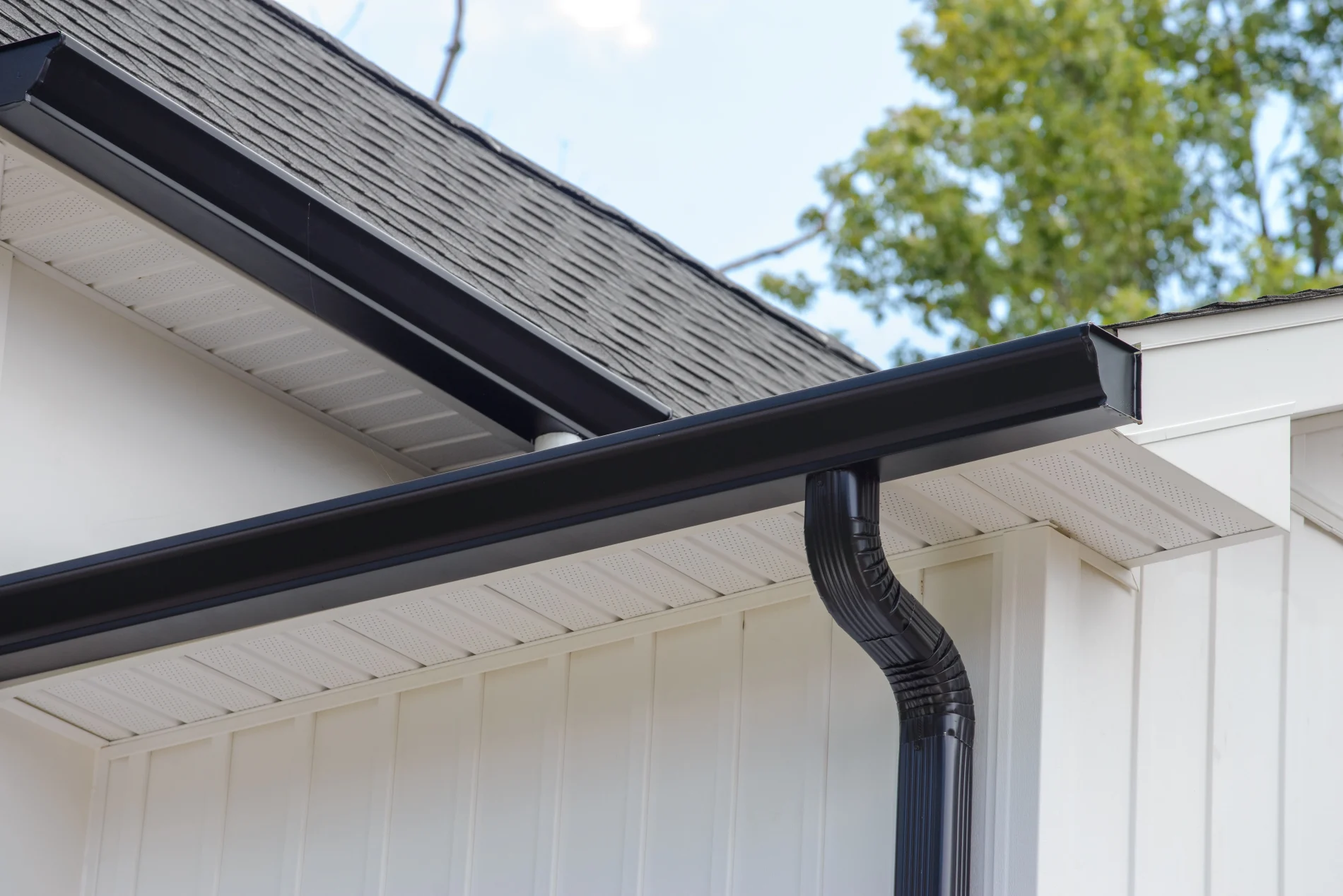 A black gutter system mounted to the edge of a gray-shingled roof on a home with white siding.