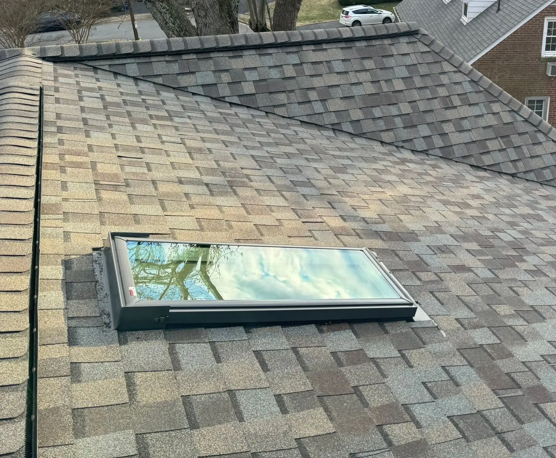 A close-up of a section of a roof’s slope that includes a rectangular skylight. The roof is covered in brown and tan shingles.