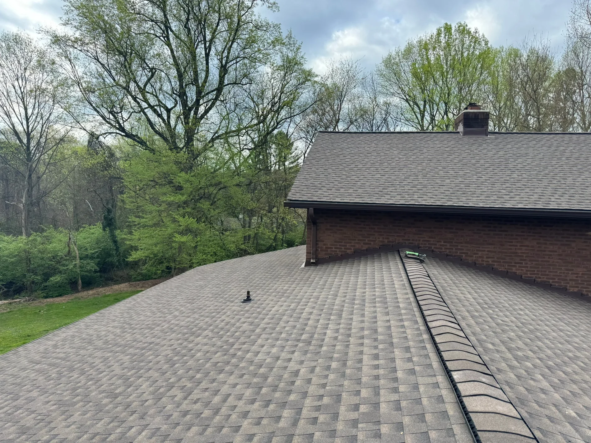 A photo taken at one end of a roof that has brown and black shingles. The home’s exterior is red brick, and there are multiple green trees seen in the distance.