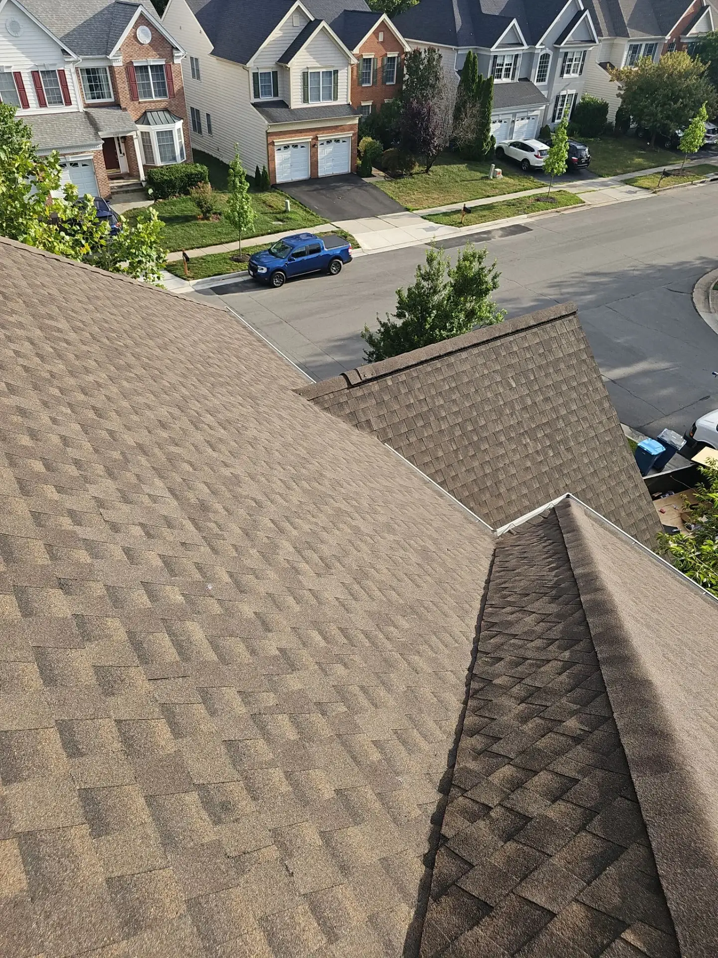 A photo taken from the top of a roof’s slope, looking down toward the street. The roof has multiple sections, and is covered in brown asphalt shingles.